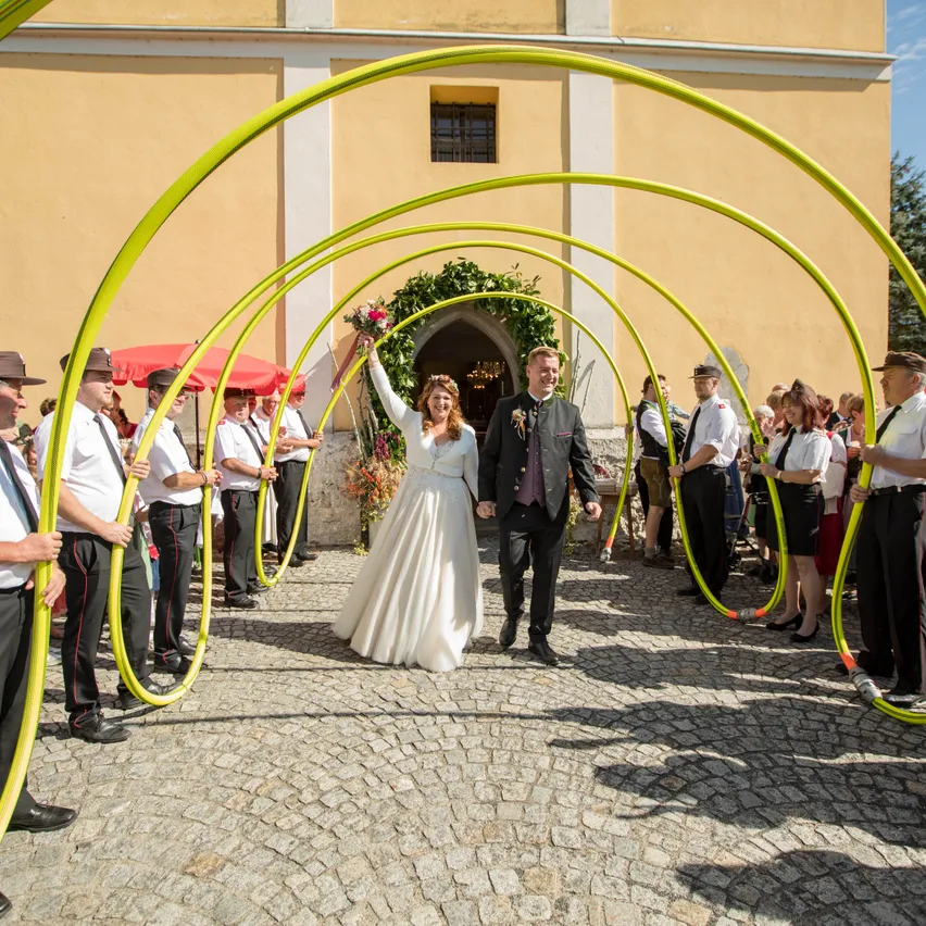 Ein frisch verheiratetes Paar geht durch einen gelben Bogen, umgeben von Zuschauern in traditioneller Kleidung.