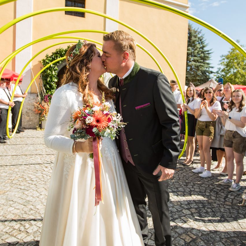 Ein frisch verheiratetes Paar küsst sich unter einem gelben Bogen, umgeben von einer Menge Schaulustiger.