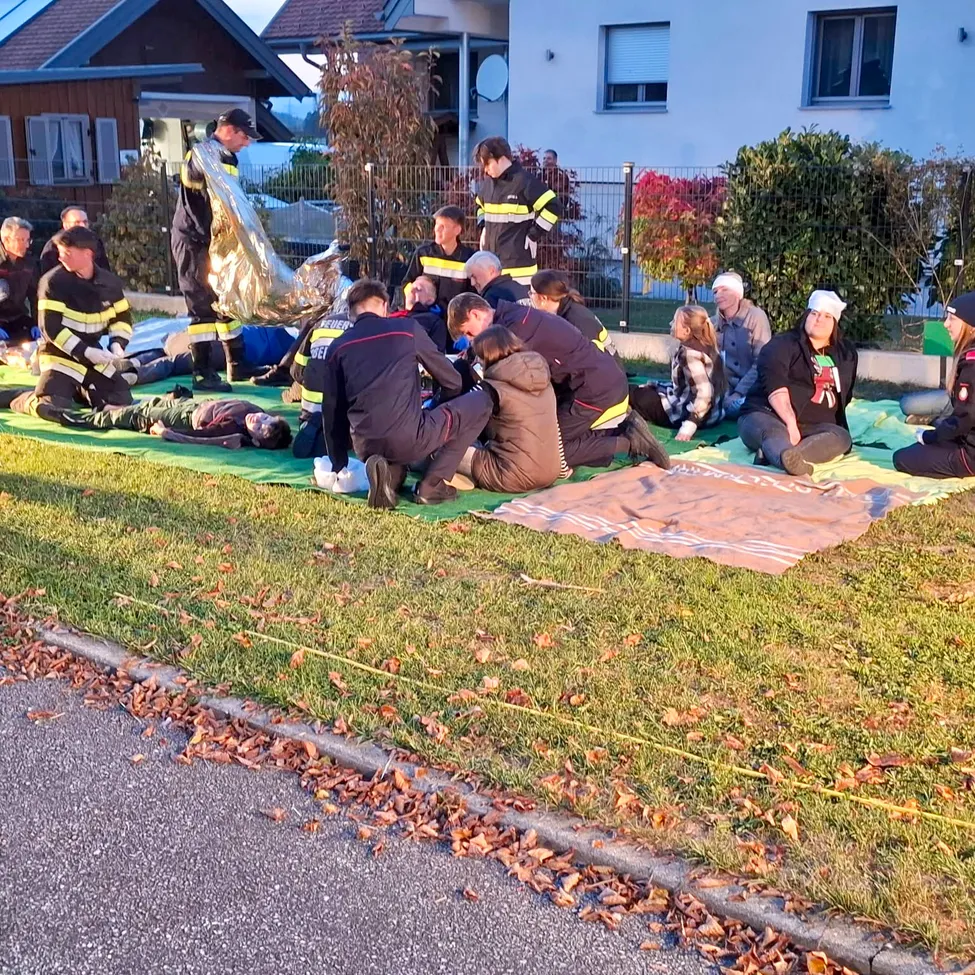 Eine Gruppe von Feuerwehrleuten kümmert sich um verletzte Menschen auf dem Boden vor einem Haus.