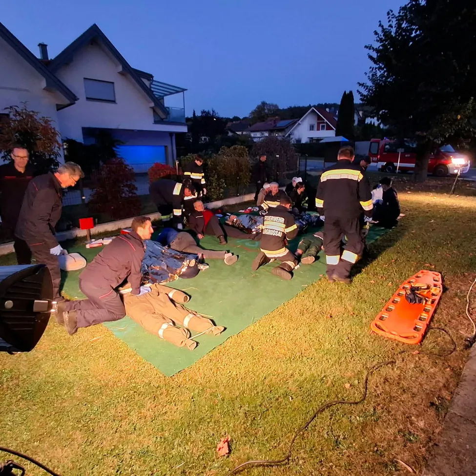 Eine Gruppe von Menschen, wahrscheinlich Feuerwehrleute, versammelt sich um eine grüne Matte auf einem Rasen. Einige sitzen, andere stehen und sie tragen alle Uniformen.