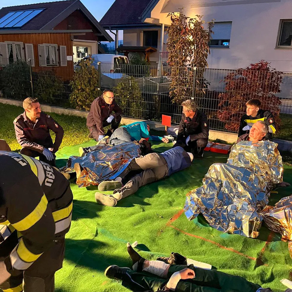 Eine Gruppe von Feuerwehrleuten und Rettungskräften kümmert sich um verletzte Menschen auf einem grünen Teppich im Hof.