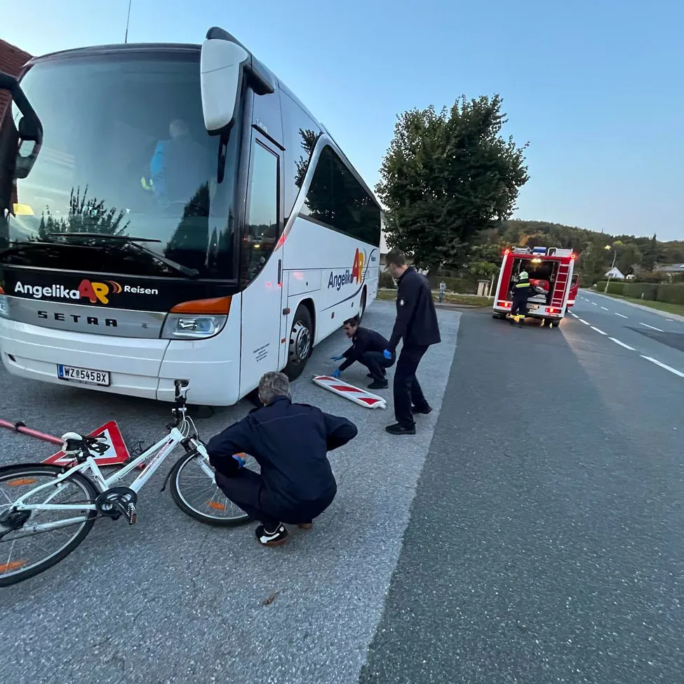 Ein Bus mit der Aufschrift Angelika Reisen ist am Straßenrand geparkt. Ein Mann repariert ein Fahrrad, während ein anderer Mann in der Nähe steht. Ein Feuerwehrwagen ist am Straßenrand geparkt.