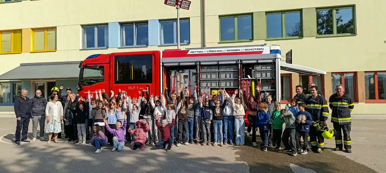 Eine Gruppe von Kindern und Erwachsenen posiert für ein Foto vor einem roten Feuerwehrauto. Das Feuerwehrauto steht vor einem Gebäude mit vielen Fenstern.