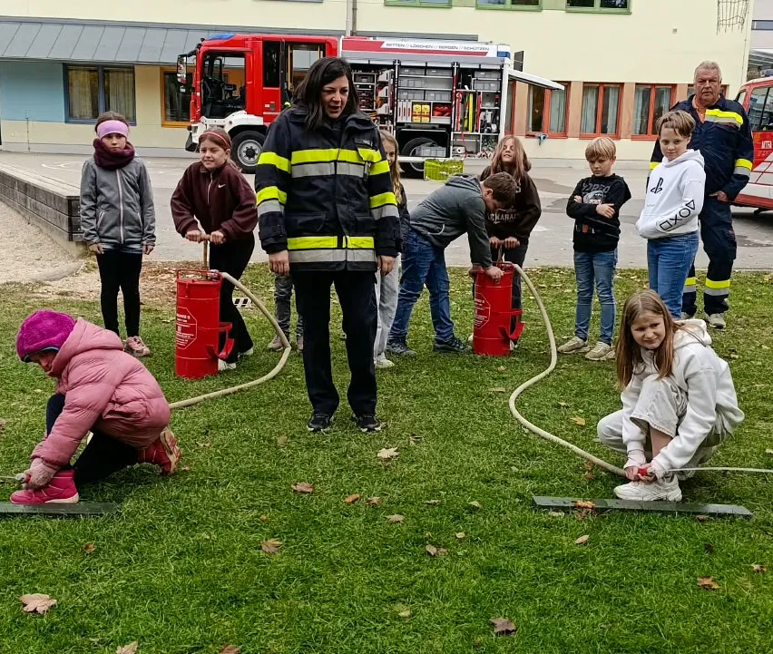 Eine Gruppe von Kindern und Erwachsenen versammelt sich auf einem Rasenfeld und nimmt an einer Brandschutzvorführung teil. Sie füllen Eimer mit Wasser aus Schläuchen, die mit Feuerhydranten verbunden sind.