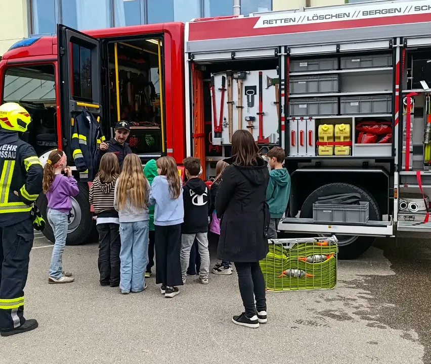 Eine Gruppe von Kindern und Erwachsenen steht vor einem Feuerwehrauto. Ein Feuerwehrmann steht neben dem Auto und scheint den Kindern etwas zu erklären.