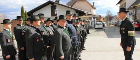 A group of formally dressed men stand in a row, wearing hats and green sashes. They are outside, in front of a building with a chimney. Cars are parked on the side of the road.