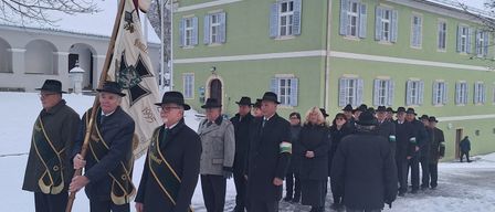 Eine Gruppe formell gekleideter Männer und Frauen steht in einem verschneiten Bereich und hält eine Fahne. Sie stehen vor einem Gebäude mit Schornstein und Schnee auf dem Dach.