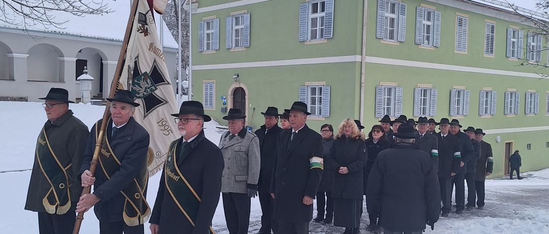 Eine Gruppe formell gekleideter Männer und Frauen steht in einem verschneiten Bereich und hält eine Fahne. Sie stehen vor einem Gebäude mit Schornstein und Schnee auf dem Dach.