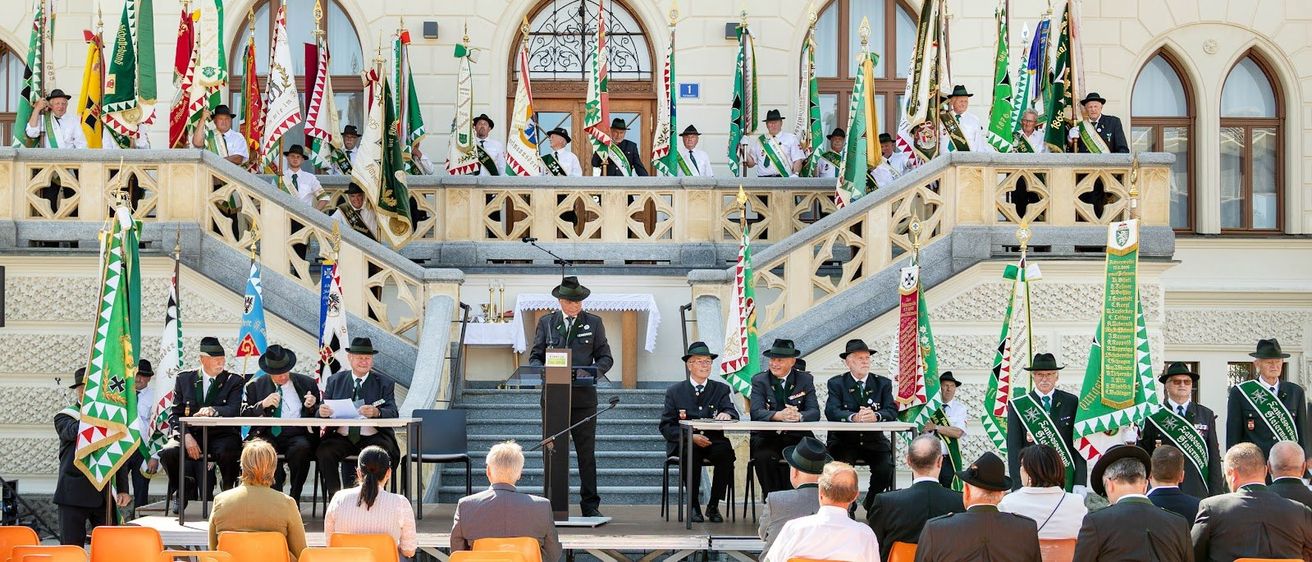 Eine Versammlung von Personen, einschließlich uniformierter Personen, findet auf den Stufen eines Gebäudes mit der Aufschrift Rathaus statt. Ein Podium mit Mikrofon steht in der Mitte, mit Personen auf beiden Seiten. Fahnen sind an den Seiten des Gebäudes platziert.