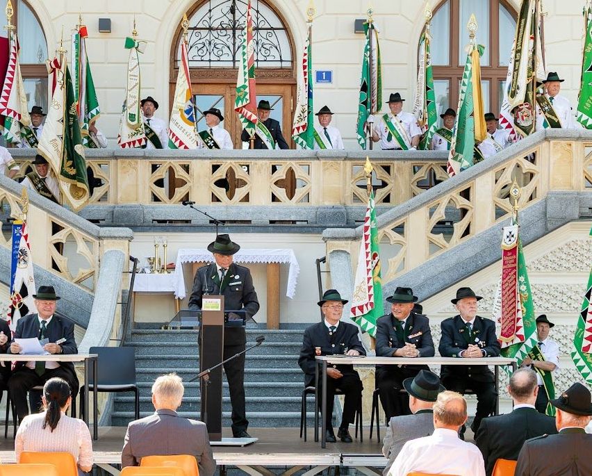 Eine Versammlung von Personen, einschließlich uniformierter Personen, findet auf den Stufen eines Gebäudes mit der Aufschrift Rathaus statt. Ein Podium mit Mikrofon steht in der Mitte, mit Personen auf beiden Seiten. Fahnen sind an den Seiten des Gebäudes platziert.