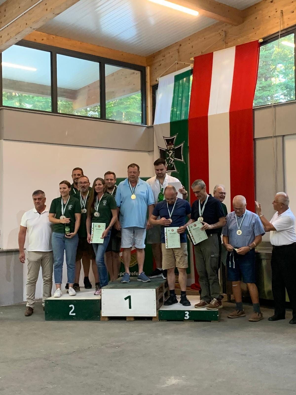 A group of people stand on a podium with medals around their necks, holding certificates. Behind them, flags hang on the wall.