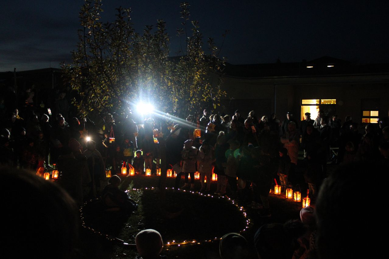 At night, a group of people gather around a circle of lit lanterns in an outdoor area. They stand in the dark, illuminated by the lanterns and a bright light. Trees surround the area.