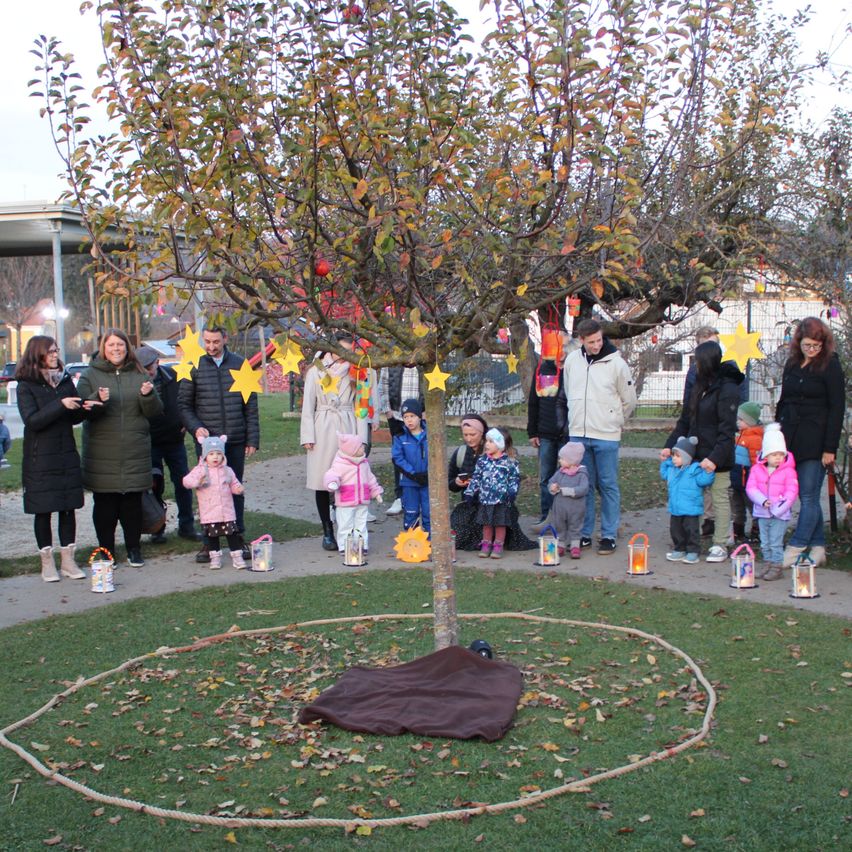 A group of people stand around a decorated tree with yellow stars. Some hold lanterns. The ground is circled with rope.