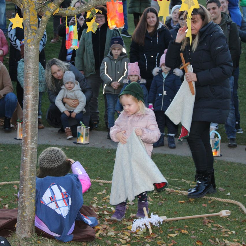 A group of people, including adults and children, are gathered around a tree. Some hold colorful lanterns. A woman holds a child's hand, while another child holds a cloth.