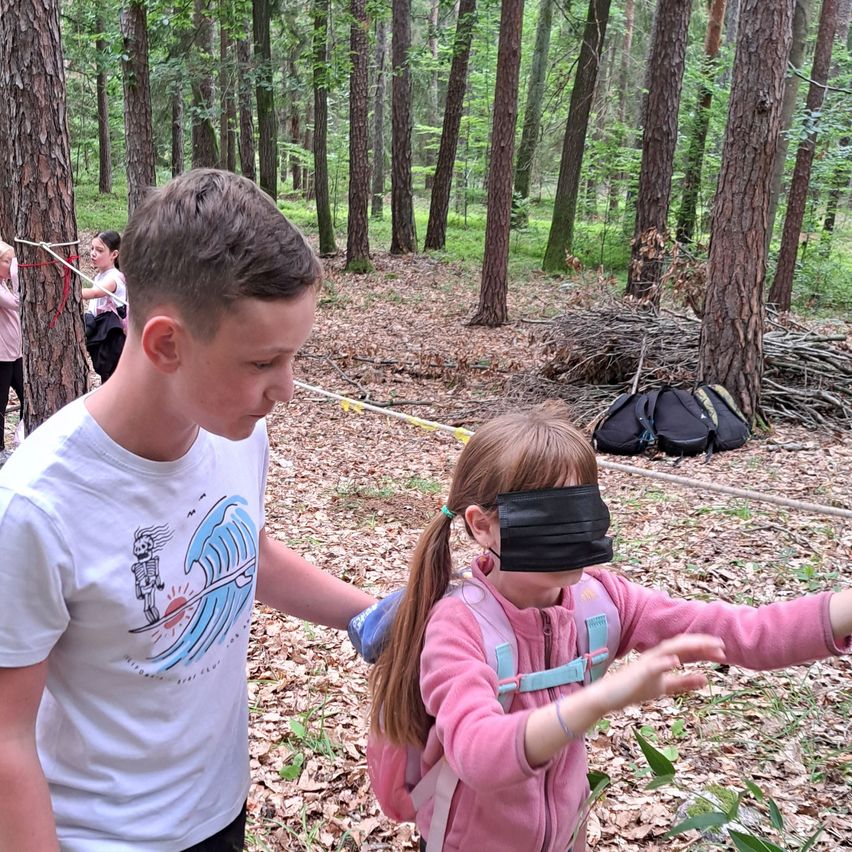 Ein Junge und ein Mädchen sind mit verbundenen Augen und erkunden einen Wald. Die Hände des Mädchens sind ausgestreckt. Sie werden von einem Jungen geführt. Im Hintergrund bindet ein anderes Mädchen ein Seil um einen Baum.