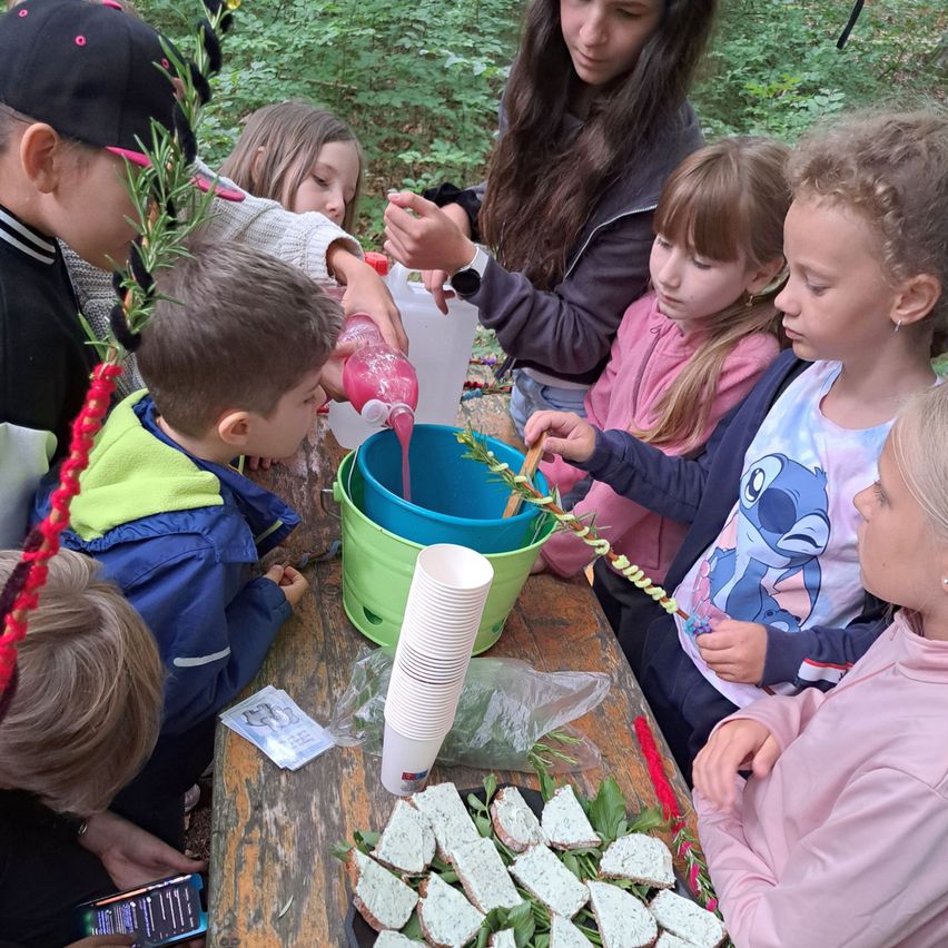 Eine Gruppe von Kindern und einer Frau versammelt sich um einen Tisch im Wald und gießt rosa Flüssigkeit in einen Eimer. Der Tisch ist mit Tellern mit Essen und Bechern bestückt.