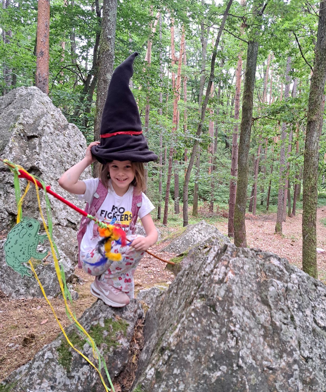 Ein kleines Mädchen in einem Hexenhut steht auf einem Felsen im Wald. Sie hält eine Spielzeugangel und lächelt.