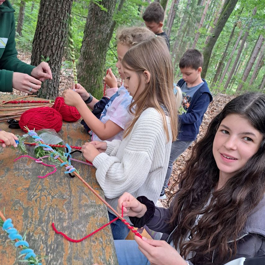 Eine Gruppe von Kindern in einer Waldumgebung arbeitet an einem Bastelprojekt. Ein Mädchen lächelt in die Kamera, während andere sich auf das Auffädeln von buntem Garn konzentrieren.