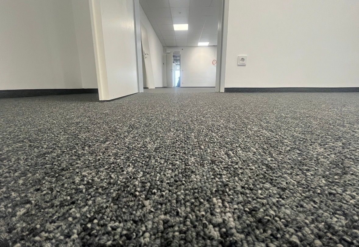 An empty office hallway with gray carpet, white walls, and two open doors. A power outlet and sign are visible on one wall.