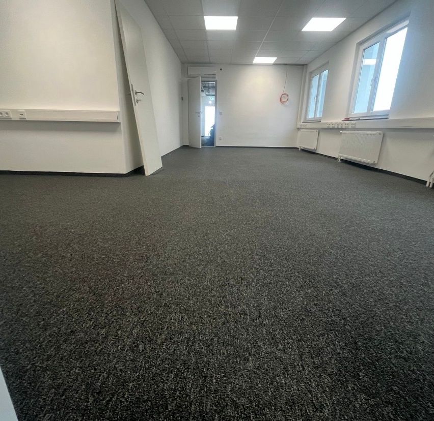 An empty office room with white walls, gray carpet, three windows, and two radiators under the windows. The room has a door open to the left and another door at the back.