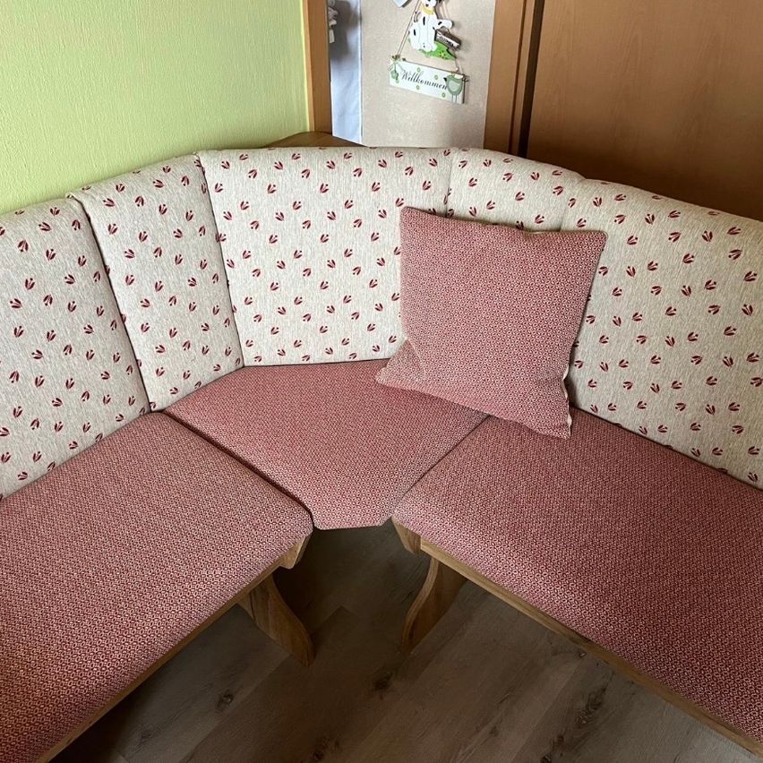 A corner sofa with a light pink cushion and a red leaf pattern on the upholstery. A small pillow rests on the cushion.