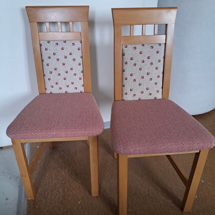 Two wooden chairs with red floral patterns on the backrests and pink cushioned seats are placed side by side against a plain white wall.