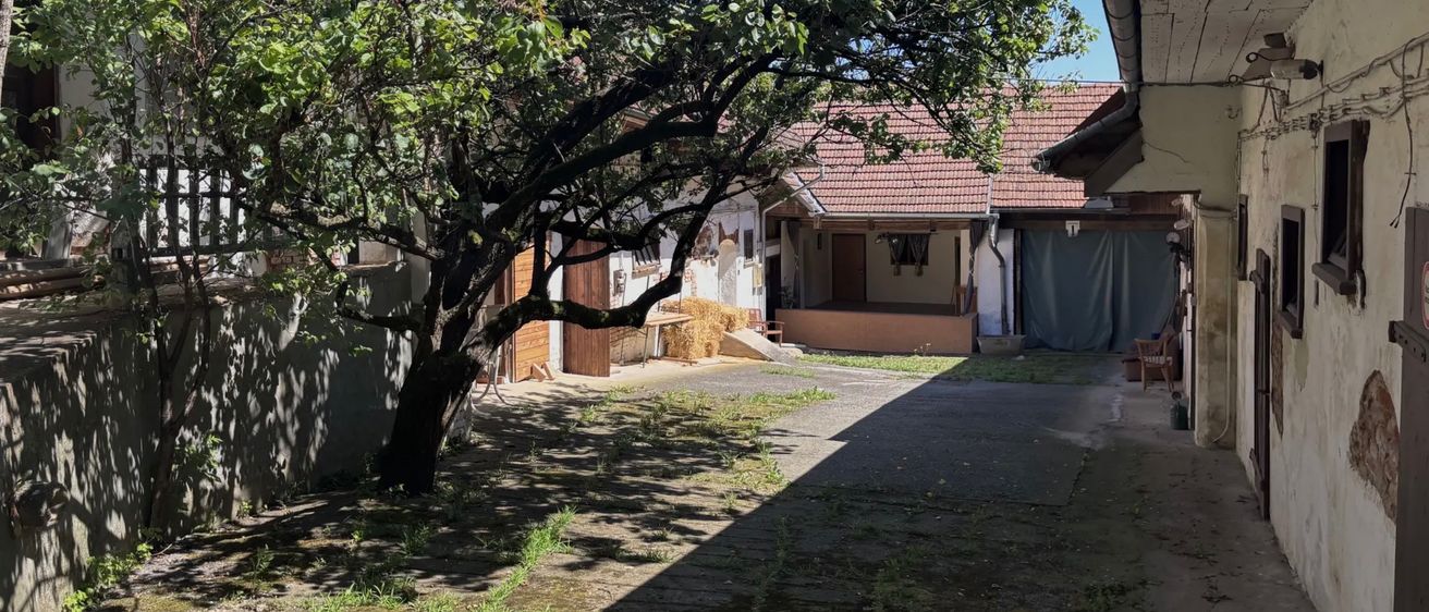 Ein Haus mit rotem Dach und einem Baum davor. Im Hof gibt es einige Bäume, eine Bank und einen Heuhaufen. Das Haus hat eine Holztür und ein Fenster.