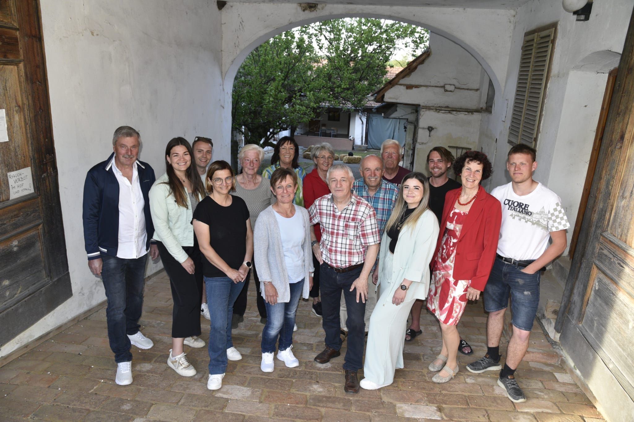Eine Gruppe von Menschen steht zusammen auf einem gepflasterten Weg und lächelt für ein Foto. Sie tragen Freizeitkleidung, einige mit Brillen und Accessoires. Dahinter ist ein Gebäude mit einem Bogen und einem Baum zu sehen.