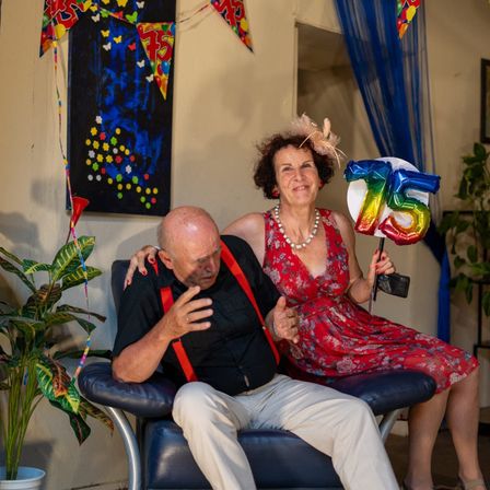 Ein älteres Paar sitzt gemeinsam auf einem Stuhl und lächelt für ein Foto. Eine Frau hält einen 75. Geburtstagsballon, und sie sind von Dekorationen und einer Topfpflanze umgeben.