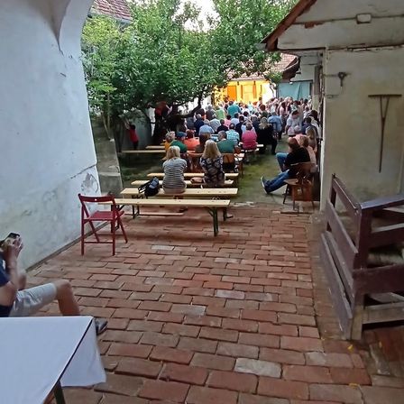 Ein Innenhof voller Menschen, einige sitzen auf Bänken, andere stehen, unter klarem Himmel. Bäume und ein Gebäude im Hintergrund.