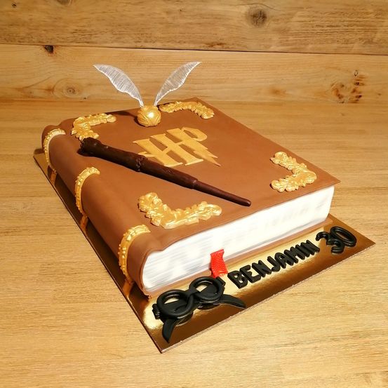 Ein Kuchen in Form eines Buches mit dem Hogwarts-Wappen und einem Zauberstab darauf, mit dem Namen Benjamin und der Nummer 30 darunter. Er hat goldene Dekorationen und einen hölzernen Hintergrund.