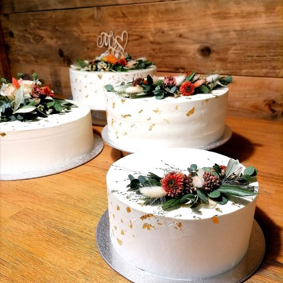 Vier weiße Hochzeitstorten mit Goldblättern und Blumendekorationen stehen auf einem Holztisch. Jeder Kuchen hat eine einzigartige Blumenanordnung. Dahinter ist eine Holzwand.