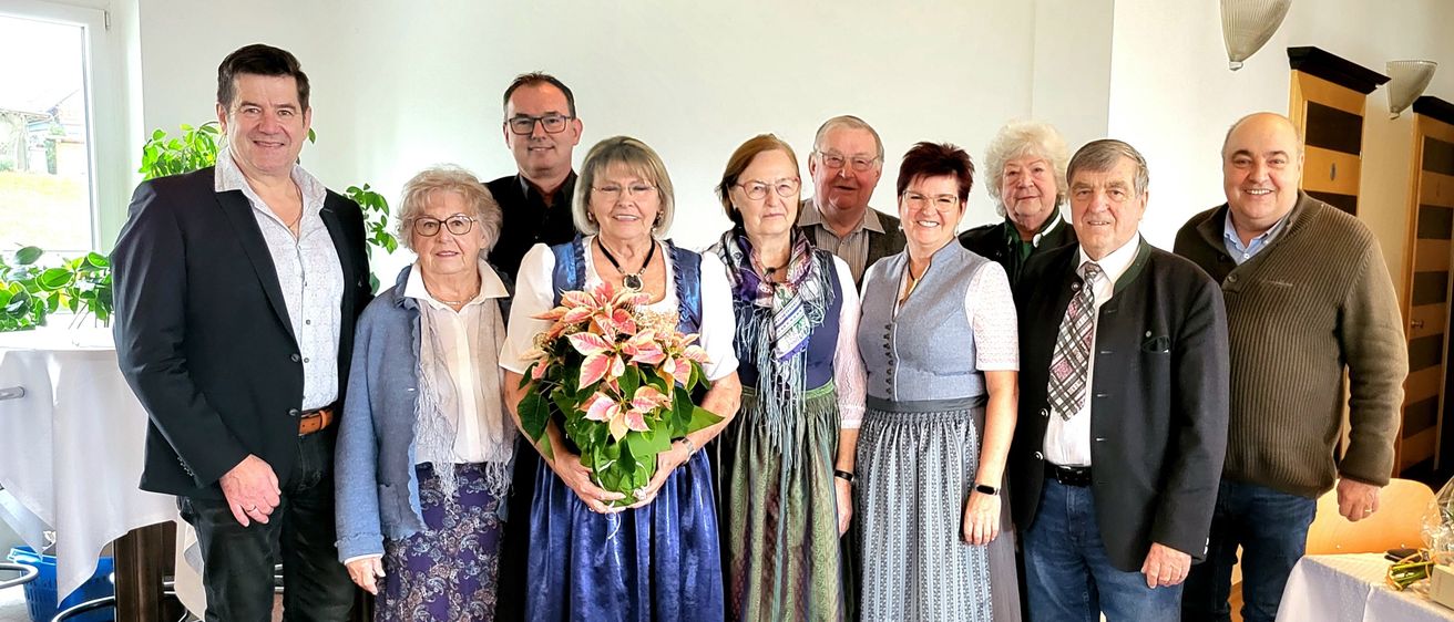 Eine Gruppe älterer Erwachsener, einige in traditioneller Kleidung, versammeln sich für ein Foto. Sie stehen eng beieinander und lächeln. Eine Frau hält einen pinken Blumenstrauß.