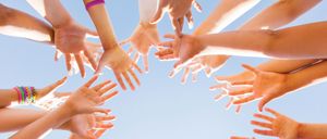 Mehrere Hände von Menschen bilden einen Kreis mit nach oben gerichteten Handflächen. Das Bild ist vor einem blauen Himmel aufgenommen.