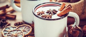 Zwei weiße Tassen mit roter Flüssigkeit, verziert mit Sternanis und Zimtstangen, auf einer Holzoberfläche mit getrockneten Orangenscheiben.