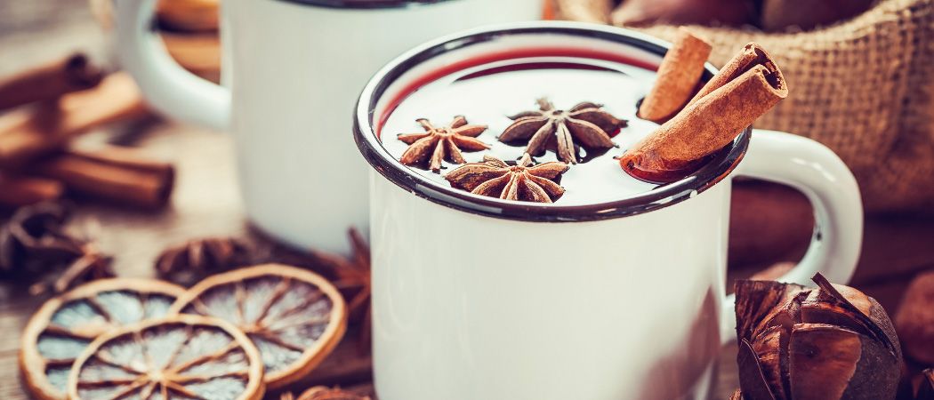 Zwei weiße Tassen mit roter Flüssigkeit, verziert mit Sternanis und Zimtstangen, auf einer Holzoberfläche mit getrockneten Orangenscheiben.