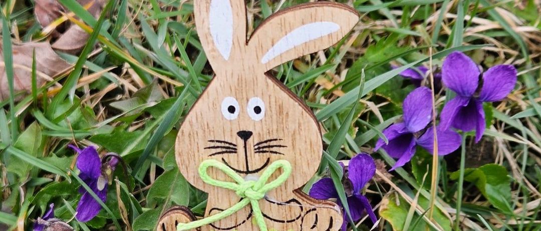 Ein hölzerner Osterhase mit einer grünen Schleife um den Hals liegt auf Gras mit violetten Blumen.