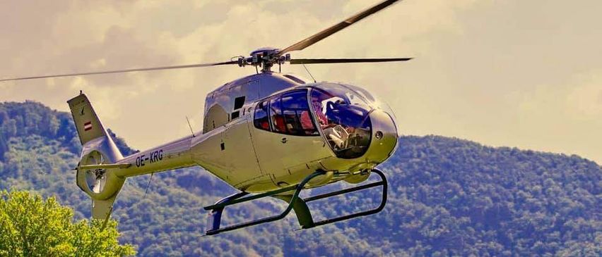Ein Hubschrauber mit der Registrierung OE-KRG fliegt über einer bewaldeten Berglandschaft mit bewölktem Himmel.