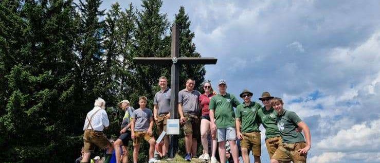 Eine Gruppe von Menschen steht auf einem Berggipfel, einige in grünen Shirts und Shorts, andere in traditioneller bayerischer Kleidung, alle posieren für ein Foto vor einem Holzkreuz.