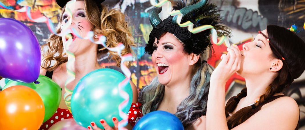 Drei Frauen in festlicher Kleidung, die bunte Ballons halten, lachen und genießen eine ausgelassene Feier, möglicherweise auf einer Party.