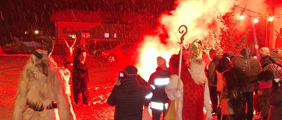Eine Menschenmenge versammelt sich um ein Feuer mit dem Weihnachtsmann und Krampus bei einer verschneiten Nacht. Ein Mann in einem weißen Kostüm hält einen Stab.