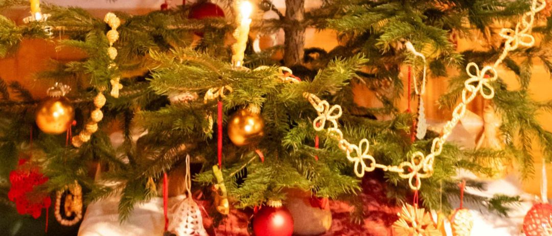 Ein Weihnachtsbaum mit roten und goldenen Ornamenten, beleuchtet mit Kerzen, steht in einem gemütlichen Raum mit Holzwänden und Fenstern.