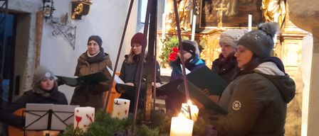 Eine Gruppe von Menschen versammelt sich in einer Kirche und singt um einen dekorierten Kerzenständer herum. Sie tragen alle Winterkleidung und Mützen. Eine Gitarre steht auf einem Notenständer auf der linken Seite.