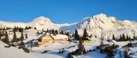 Eine Holzhütte mit schneebedecktem Dach steht in einer verschneiten Landschaft mit Bergen im Hintergrund.