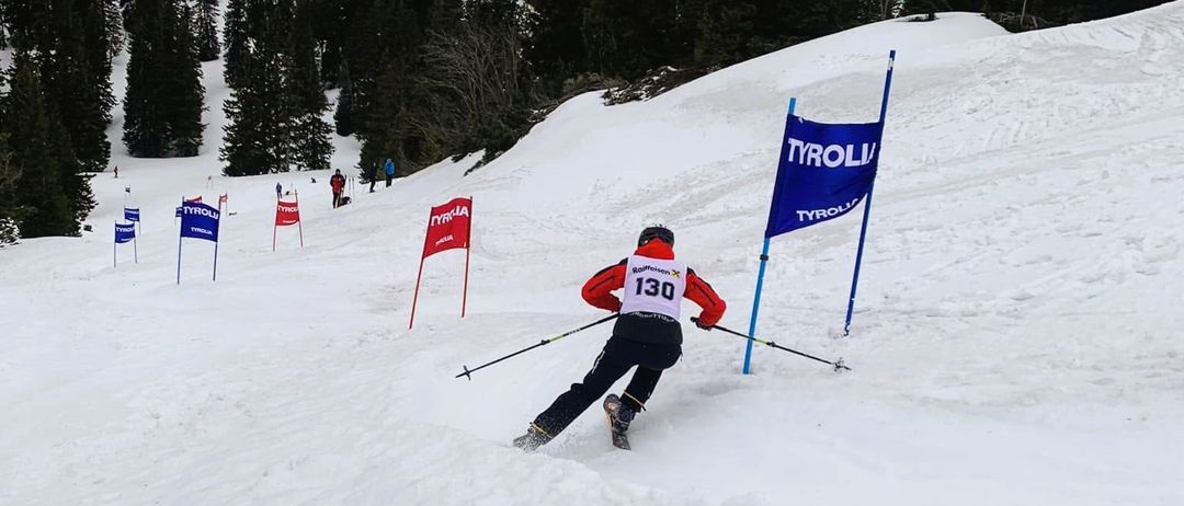Ein Skifahrer in einem roten und weißen Outfit mit der Nummer 130 fährt einen schneebedeckten Hang hinunter, an dem mehrere rote Flaggen und eine blaue Flagge mit der Aufschrift Tirol angebracht sind.