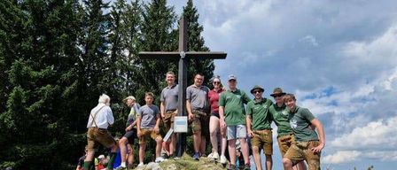 Eine Gruppe von Menschen, wahrscheinlich auf einer Wanderung, posiert für ein Foto mit einem Kreuz auf einem Hügel, umgeben von Bäumen und einem bewölkten Himmel.