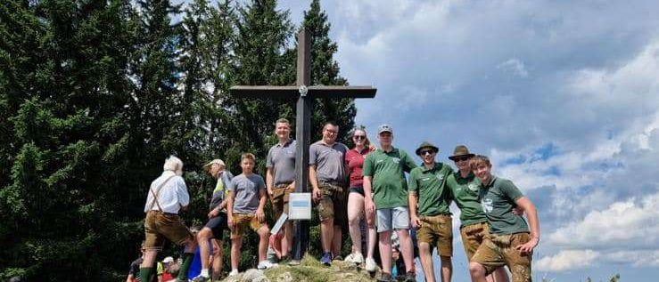Eine Gruppe von Menschen, wahrscheinlich auf einer Wanderung, posiert für ein Foto mit einem Kreuz auf einem Hügel, umgeben von Bäumen und einem bewölkten Himmel.