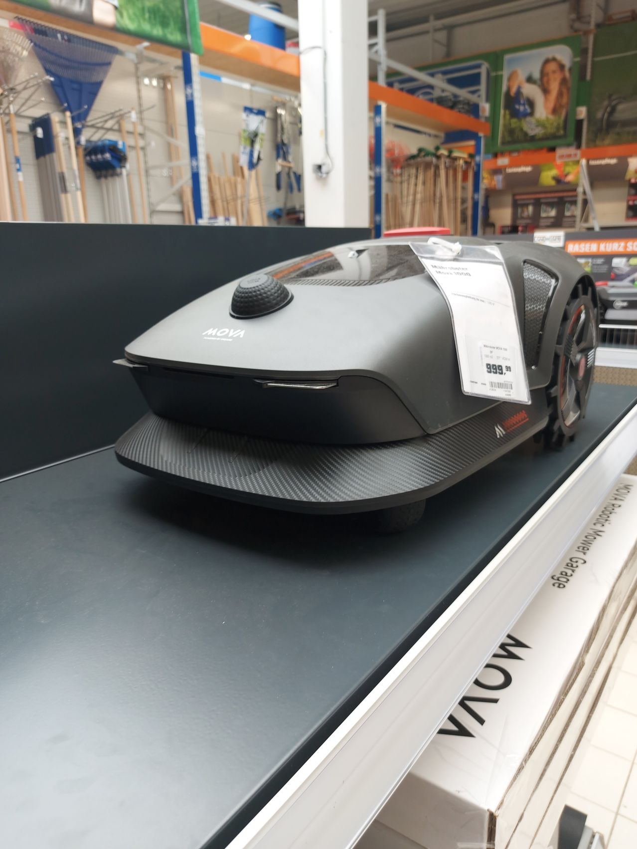 Ein schwarz-grauer robotergesteuerter Rasenmäher wird in einem Geschäft auf einem Tisch ausgestellt. Der Mähroboter steht auf einer schwarzen Oberfläche mit einem Preisschild von 999 Euro.