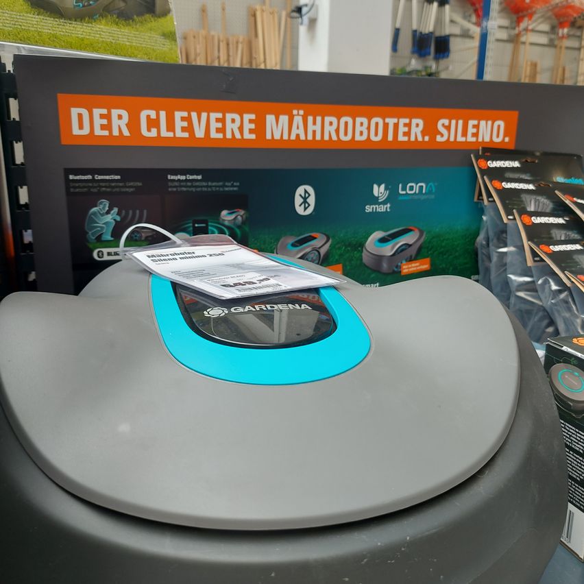 Ein grauer und blauer Rasenmähroboter der Marke Gardena, ausgestellt in einem Geschäft. Die Anzeige dahinter bewirbt das Produkt mit deutschem Text.