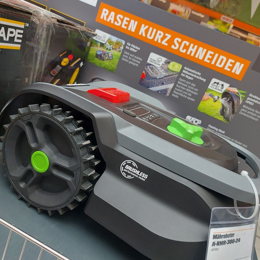 Ein schwarzer und grauer Roboter-Rasenmäher wird in einem Geschäft ausgestellt, an dem ein Schild mit der Aufschrift 'Maehrobot A-RMR-300-24' hängt. Der Mähroboter verfügt über ein großes Rad und einen grünen Knopf. Ein Schild im Hintergrund trägt die Aufschrift 'RASEN KURZ SCHNEIDEN'.